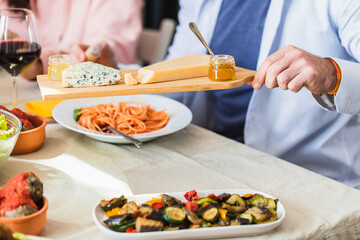 Man holding wooden tray with cheese and honey at dinner table with friends