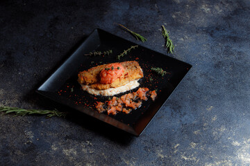 Grilled salmon steak served with fresh tomatoes, pepper seeds, rosemary, thyme on black plate on dark table.