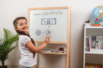 Little girl stands near a white board, smiles and shows how she successfully solves mathematical problems using banknotes and coins