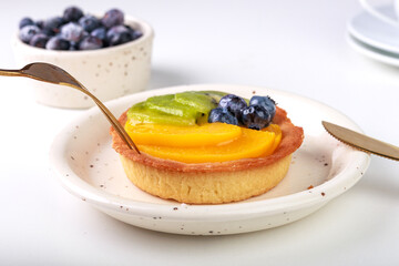 shortbread basket cake with cream, blueberries, peaches and kiwi on a plate on a white background, golden cutlery