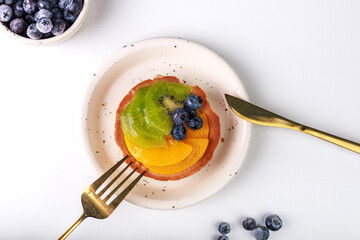 shortbread basket cake with cream, blueberries, peaches and kiwi on a plate on a white background, golden cutlery