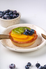 shortbread basket cake with cream, blueberries, peaches and kiwi on a plate on a white background, golden cutlery
