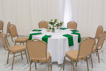 Wine glasses, silver tableware and green napkins on round tables covered with white tablecloth.