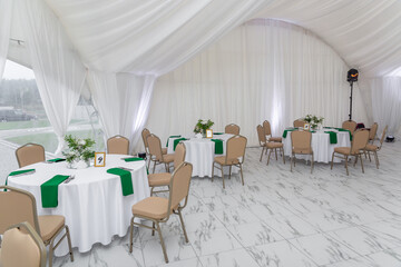 Wine glasses, silver tableware and green napkins on round tables covered with white tablecloth.