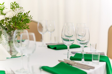 Close up of wine glasses, silver tableware and green napkins on round tables covered with white tablecloth.