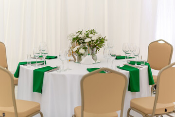 Wine glasses, silver tableware and green napkins on round tables covered with white tablecloth.