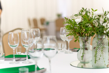 Close up of wine glasses, silver tableware and green napkins on round tables covered with white tablecloth.