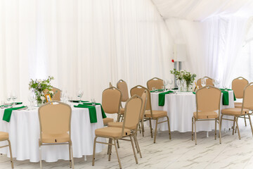 Wine glasses, silver tableware and green napkins on round tables covered with white tablecloth.