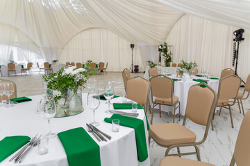 Wine glasses, silver tableware and green napkins on round tables covered with white tablecloth.