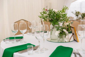 Close up of wine glasses, silver tableware and green napkins on round tables covered with white tablecloth.