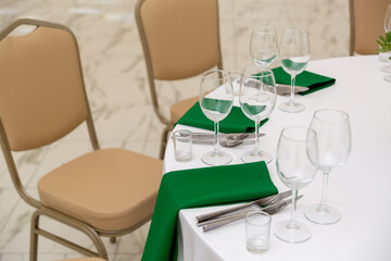 Close up of wine glasses, silver tableware and green napkins on round tables covered with white tablecloth.