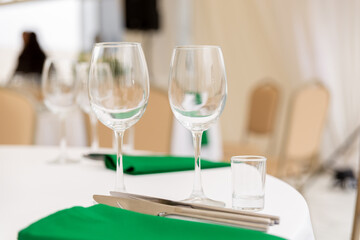 Close up of wine glasses, silver tableware and green napkins on round tables covered with white tablecloth.