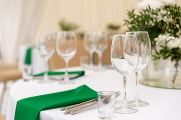 Close up of wine glasses, silver tableware and green napkins on round tables covered with white tablecloth.