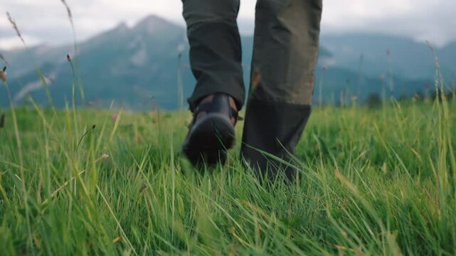 Rear back view gimbal shot male feet legs stepping on grassy meadow hill overlooking mountains in sunrise or sunset light. Man wearing hiking boots walks on clover grass. Male hiker explorer