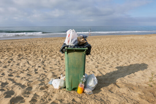 An overflowing green garbage bin on a sandy beach with scattered trash bags around it, set against a cloudy oceanfront background, highlighting beach maintenance and environmental concerns