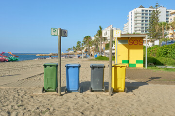 Four recycling bins on the sandy beach of Torre Bermeja in Benalmadena, Malaga, Andalusia, Spain. Next to a lifeguard station, framed by a coastal cityscape and a clear blue sky