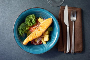 Top view of grilled salmon fillet served with steamed broccoli and roasted vegetables in a turquoise ceramic plate with a fork and knife, and brown napkin on dark background