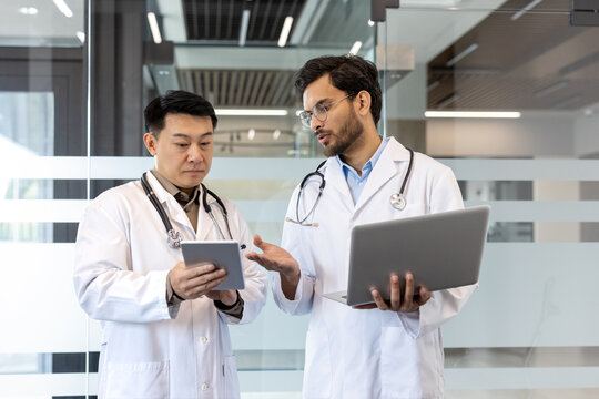 Two doctors discussing medical information using tablet and laptop, showcasing collaboration in hospital. Stethoscopes indicate professional medical setting, emphasizing teamwork and technology use.