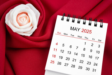 May 2025 desk calendar and pink rose on red textile.