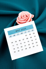May 2025 calendar page and pink rose on green textile.