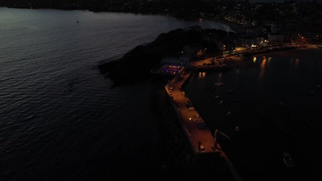 Aerial view of Panx&oacute;n harbor during blue hour. Vibrant summer party takes place on a crowded terrace. Camera moves backward, revealing Panx&oacute;n's harbour, and the illuminated pier with anchored boats. 