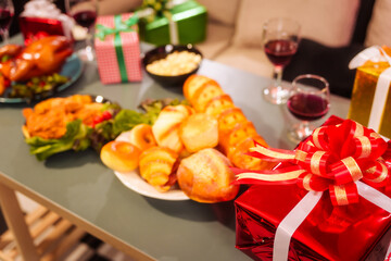 The cozy Christmas party setup features variety of beautifully wrapped gift boxes on table, alongside festive food dishes fried chicken, grilled chicken, salad, popcorn, and bread in living room.