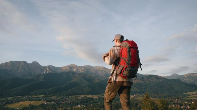 Back side view following shot of man with red backpack walking down the hill overlooking epic mountains lit with soft sunrise light. Male backpacker explorer hiking, sunny summer morning