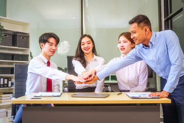 In a collaborative office setting, two men and two women, including a boss and employees, join hands and raise their hands in unity, celebrating teamwork and achieving a business target together.