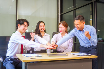 In a collaborative office setting, two men and two women, including a boss and employees, join hands and raise their hands in unity, celebrating teamwork and achieving a business target together.