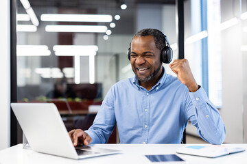 Mature African American businessman wearing headphones celebrates success at desk in modern office. Happy, motivated professional enjoying achievement. Laptop in foreground signifies work and triumph