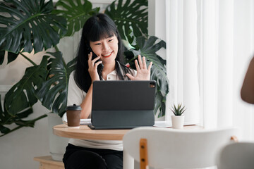 Smiling woman on a video call using a laptop and smartphone, sitting at a table with plants and coffee in a bright home office.