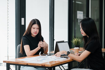 Two young women working together in a contemporary office setting, discussing documents and using laptops.