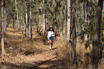 Obraz premium Latin woman, young, beautiful, dark-haired, with curly hair, with a white shirt and top, denim skirt and hat, walking alone and calmly through a eucalyptus forest. Concept peace, relaxation.