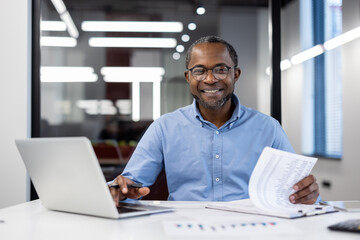 Obraz premium Mature african american businessman smiling confidently working on laptop in modern office. Business setting with important documents showcasing success and professionalism.