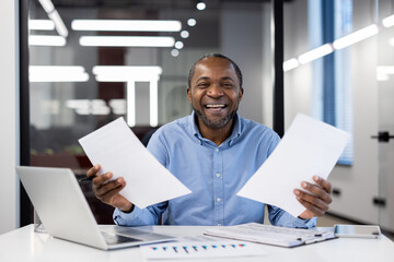 Mature african american businessman smiling confidently, holding papers in a modern office setting. Represents success, positivity, and professionalism in workplace environment.