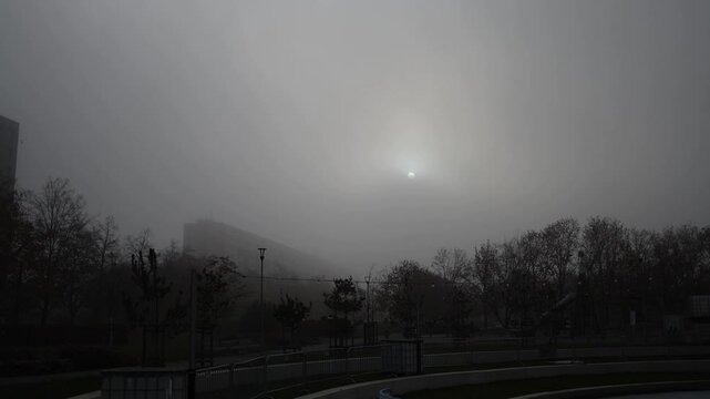 A foggy morning scene with the sun barely visible through thick mist, creating an eerie and calm atmosphere. The outlines of trees and distant buildings fade into the gray haze, adding a sense of soli