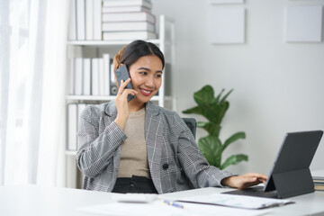 Businesswoman in a modern office talking on the phone while working on a laptop, smiling, and handling paperwork efficiently.