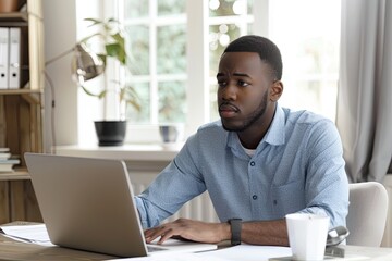 Fototapeta premium man in a blue shirt looks focused as he works on his laptop in a bright, organized home office