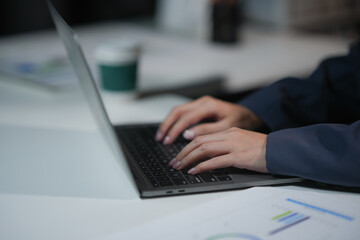 Close-up of hands typing on a laptop keyboard in an office setting, with charts and a coffee cup in the background.