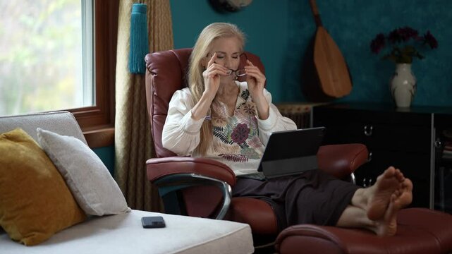 Using glasses to see tablet computer, a mature woman with blonde hair sits in a brown leather recliner. Her feet are propped up on the footrest and she has a relaxed smile. 
