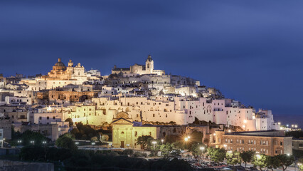 Naklejka premium Ancient Italian town on hill top illuminated at night against blurred cloudy sky, Ostuni, Puglia, Italy