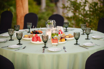 Elegant Round Table Arranged For Outdoor Gathering With Fruits, Pastries, And Refreshments In Natural Light Surrounded By Greenery.