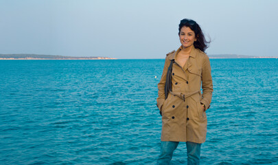 Smiling woman in a trench coat standing by the sea with a scenic view, wind blowing her hair,...