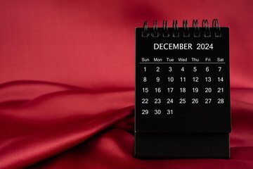 Black desk calendar for December 2024 on a red satin or silk textile background.