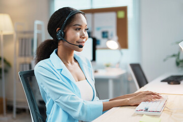 Friendly female call center agent wearing headset assisting client online, typing on keyboard,...