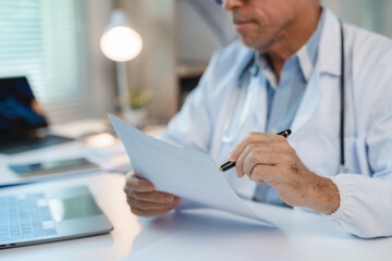 Senior doctor reviewing patient's medical history, holding documents and pen, sitting at desk with...