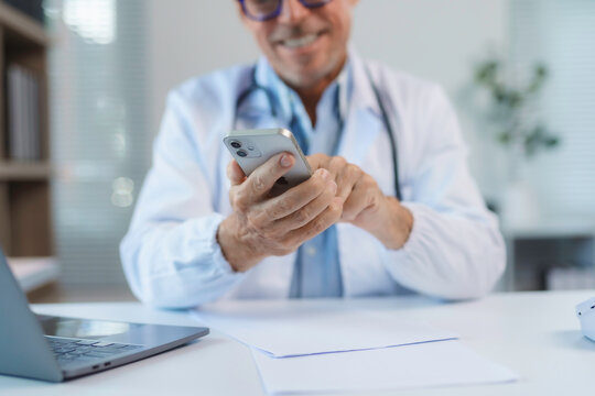 Smiling doctor using smartphone in medical office with laptop and documents on desk