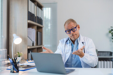 Mature doctor wearing lab coat and stethoscope having a video conference on his laptop in medical office