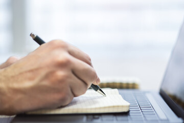 Tight shot of a man's hand penning in a notepad on a chic laptop, background blurred