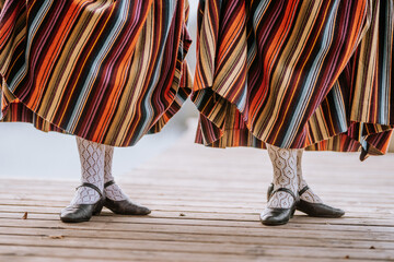 Close-up of two women’s legs wearing colorful striped skirts, lace socks, and black shoes, showcasing traditional attire on a wooden platform.